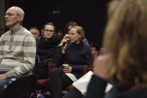 Jedna z kobiet siedzących na publiczności trzyma w ręku mikrofon przy ustach. Po prawej stronie fotografii jest kobieta, która ukazana jest tyłem.