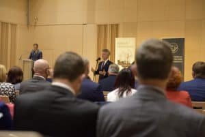 Grupa osób siedzi na krzesłach w sali. Na scenie przemawia mężczyzna w garniturze. Osoby siedzące są tyłem do obiektywu.