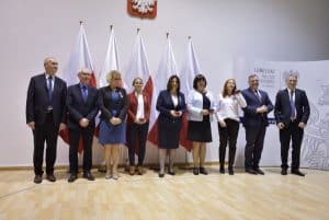 Kobiety i mężczyźni pozują do zdjęcia. Jest ich dziewięcioro. Za nimi stoi pięć flag Polski.