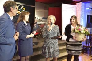 Ludzie stoją na scenie. Jedna z kobiet trzyma mikrofon w dłoniach. Jedna kwiaty.