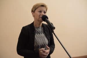 Kobieta stoi przed obiektywem i ma mikrofon blisko ust. Ręce trzyma złączone na wysokości brzucha.