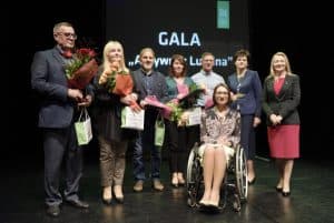 Osoby pozują do zdjęcia- trzech mężczyzn, cztery kobiety i kobieta na wózku inwalidzkim. Cztery osoby mają bukiety kwiatów i torby prezentowe. Na prezentacji za nimi jest napis: “ Gala”.