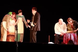 Grupa osób stoi na scenie. po prawej stronie mężczyzna i kobieta siedzą przy stoliku. Po prawej trzy kobiety i mężczyzna stoją obok siebie.