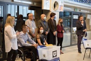 Mężczyzna przemawia do mikrofonu. Po lewej stronie stoją ludzie oraz dwie osoby na wózku. Część z nich ma głowy odwrócone w stronę mówiącego.