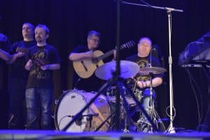 Na scenie jest kilku mężczyzn. jeden siedzi przy perkusji, a za nim stoi mężczyzna z gitarą. Obok stoi mężczyzna i trzyma instrument.