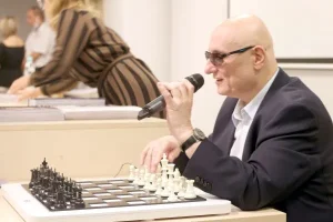 Mężczyzna trzyma mikrofon i mówi do niego. Przed nim na stole leżą szachy.