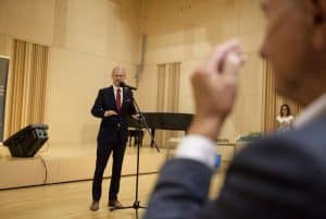 Mężczyzna w garniturze przemawia do mikrofonu. Po prawej stronie jest mężczyzna, który trzyma rękę uniesioną w górze. Jest on ucięty w obiektywie.