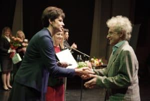 Dwie kobiety stoją naprzeciwko siebie i mają dłonie w uścisku. Kobieta za nimi trzyma otwartą teczkę i bukiet kwiatów. Dwie kobiety stoją bokiem do obiektywu.