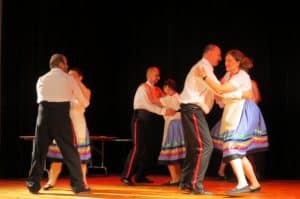 Ludzie w strojach ludowych są na scenie trzymają się za ramiona i tańcza.