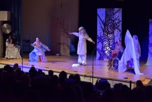 Ludzie w przebraniach są na scenie. Pada na nich niebieskie światło. W tle jest widownia.