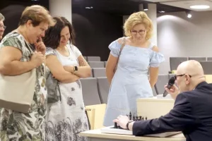 Trzy kobiety stoją przed stołem z szachami i mężczyzną. Z uwagą przyglądają się co robi mężczyzna.