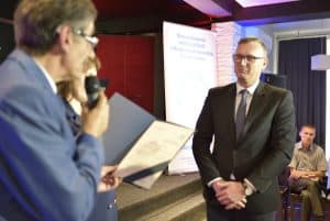 Mężczyzna ma twarz zwróconą w stronę mężczyzny, który się uśmiecha. Ma mikrofon i dyplom w dłoni.