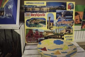 Na białej skrzyni stoi plakat ze naklejonymi zdjęciami z różnych miejsc na świecie. Przed plakatem jest drewniana miska na której są ułożone kształty wody, łódek i słońca.