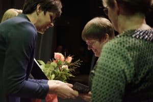 Kobieta po lewej stronie jest nachylona i trzyma w rękach pudełko a kobieta naprzeciwko trzyma ręce na pudełku. Po prawej stronie stroi konieta tyłem do obiektywu.