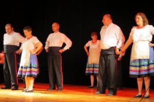 ludzie w strojach ludowych stoją na scenie i trzymają się za ręce albo mają je założone na biodra.