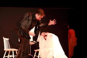 Na scenie są dwie osoby. Mężczyzna pochyla się nad kobietą, która klęczy. Trzym ona złączone dłonie na wysokości czoła i ma białą sukienkę. Za nimi stoją krzesła.