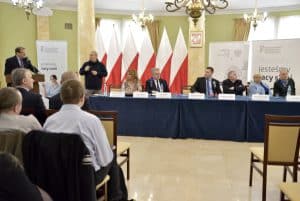 Za pulpitem stoi mężczyzna. Przy jego twarzy znajduje się mikrofon. Sześć osób – dwie kobiety i czterech mężczyzn – siedzi przy stołach przykrytych niebieskim obrusem, przed nimi na stole stoją cztery białe tabliczki z napisami. Między stołem a pulpitem stoi tłumacz języka migowego wykonujący gest rękami.