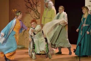 Kobieta na wózku inwalidzkim jest na scenie. Wokoło niej kobiety przebrane za księżne. Uśmechają się.