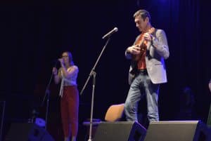 Mężczyzna stoi przy mikrofonie i trzyma gitarę. Po lewej stronie stoi kobieta, która trzyma głowę blisko mikrofonu.