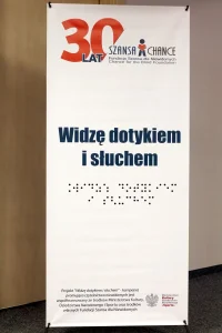 Plakat z napisem: “ Widzę dotykiem i słuchem”. Pod spodem napis braillem.