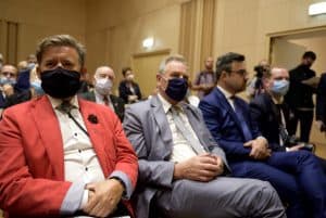 Mężczyźni w maseczkach siedzą na krzesłach. Sa ubrani w garnitury.