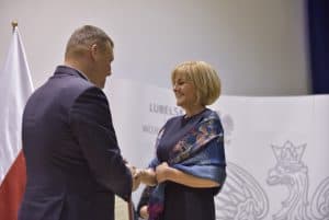 Mężczyzna i kobieta stoją naprzeciwko siebie i trzymają swoje dłonie w geście gratulacji. Kobieta uśmiecha się szeroko. Oboje są elegancko ubrani.