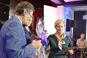 Mężczyzna stoi na scenie. W dłoni trzyma upominek zawinięte w celofan. nW tle za nim stoi kobieta, trzyma dyplom w dłoni.