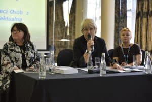 Trzy kobiety siedzą przy stole. Kobieta pośrodku trzyma mikrofon blisko twarzy. Po lewej stronie kobieta ma głowę zwróconą w bok. Kobieta po prawej stronie patrzy w obiektyw.