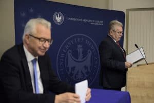 Mężczyźni stoją na scenie. Jeden stoi na mównicy. W dłoni trzyma teczkę z kartką. W tle duży baner “ Uniwersytet muzyczny w lublinie”.