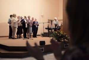 Grupa osób stoi na scenie z kartkami. Przed nimi jest mikrofon. Po prawej stronie jest niewyraźna sylwetka osoby, która trzyma ręce w górze w geście klaskania.