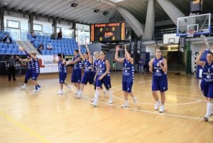 Kobiety w strojach sportowych stoją na środku boiska. Wyciągają ręce do góry.