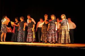 kobiety w długich spódnicach stoją na scenie. Obok jest kobieta trzymająca w dłoniach karton.