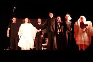 Grupa osób stoi na scenie. Mężczyzna i kobieta n a środku trzymają się za ręce. Wszyscy mają kostiumy.