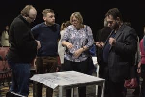 Trzech mężczyzn i jedna kobieta pochylają się nad stołem, na którym znajduje duża zapisana kartka. Dwóch mężczyzn ma pochylone głowy, natomiast jeden patrzy na kobietę. Ta jest wpatrzona w zapisy na kartce.