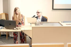 Kobieta i mężczyzna siedzą za dużym stołem. Mężczyzna mówi coś do mikrofonu a kobieta słucha.