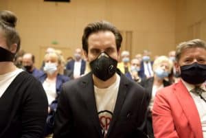 Grupa osób siedzi na widowni na krzesłach. Na pierwszym planie jest młody mężczyzna w maseczce.