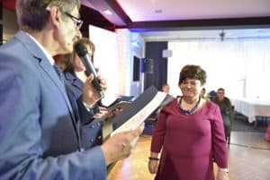 Mężczyzna trzyma w dłoni dyplom i mikrofon. Obok niego stoi kobieta. Jej twarz jest zwrócona w kierunku dyplomu.