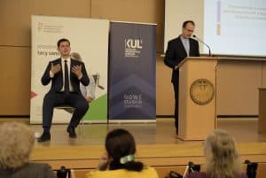 Na pierwszym planie widać trzy osoby siedzące tyłem do obiektywu. Na scenie znajdują się dwaj mężczyźni, od lewej: tłumacz języka migowego, wykonujący gest rękami, za pulpitem ksiądz, w kierunku jego twarzy skierowany jest mikrofon. Za nimi znajdują się dwie plansze od prawej: KUL „Nowe Stulecie” oraz „Wszyscy jesteśmy tacy sami”.