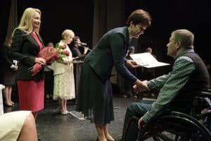 Kobieta po lewej stronie nachyla się nad mężczyzna na wózku inwalidzkim, a w ręku trzyma otwartą teczkę. Za nią stoi kobieta z kwiatami.