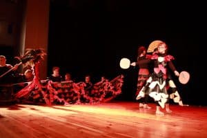 Na scenie jest grupa osób. Po prawej stronie stoi dziewczynka ubrana w sukienkę a w dłoniach ma wachlarze. Za nią na podłodze siedzą dzieci i trzymają smoka zrobionego odręcznie.