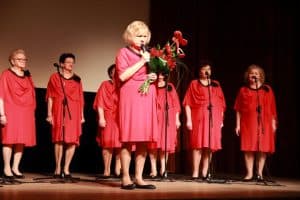 Kobieta stoi na scenie. W jednej dłoni trzyma bukiet róż, a w drugiej mikrofon. W tle inne kobiety stojące na scenie.