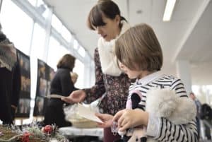 dzieci pochylają się nad stoiskiem. W rękach chłopiec trzyma kartkę. Na stole są wyroby manulane.
