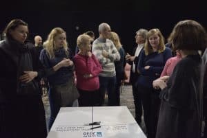 Grupa ludzi w różnym wieku stoi nad dużą kartką z tabelą, która leży na stole. Dwóch mężczyzn rozmawia ze sobą. Wszystkie kobiety z nich trzymają ręce przed sobą, czy to krzyżując je na piersiach jak również podpierając podbródek.