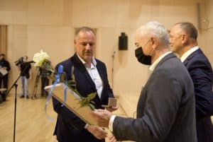 Na scenie stoi trzech mężczyzn. Starszy mężczyzna trzyma białą różę i antyramę. Drugi mężczyzna trzyma w ręce otwarte pudełeczko.