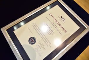 Na stole leży dyplom z napisem: “ Dyplom uznania z okazji XX-lecia Powszechnych Ubezpieczeń Zdrowotnych w Polsce dla Elżbiety Złoto”. Dalszy tekst jest zamglony.