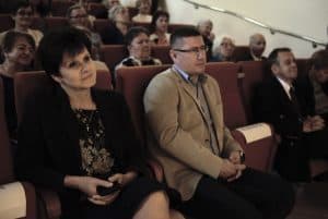 Osoby siedzą na widowni i patrzą przed siebie. Kobieta po lewej stronie trzyma w rękach komórkę i lekko się uśmiecha.