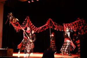 Na scenie grupa osób jest przebrana w czerwono-czarne stroje. Osoby trzymają na patykach smoka.