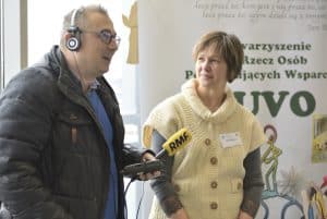 Mężczyzna trzyma w dłoni mikrofon z logiem “RMF FM”. Kobieta obok patrzy na tego mężczyznę.