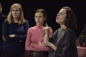 Tłumaczka języka migowego tłumaczy coś publiczności. Obok niej stoją dwie kobiety. Obie mają ręce skrzyżowane na piersiach.