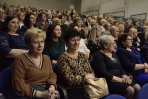 Kobiety i mężczyźni siedzą na widowni. Wszyscy są elegancko ubrani i patrzą przed siebie.